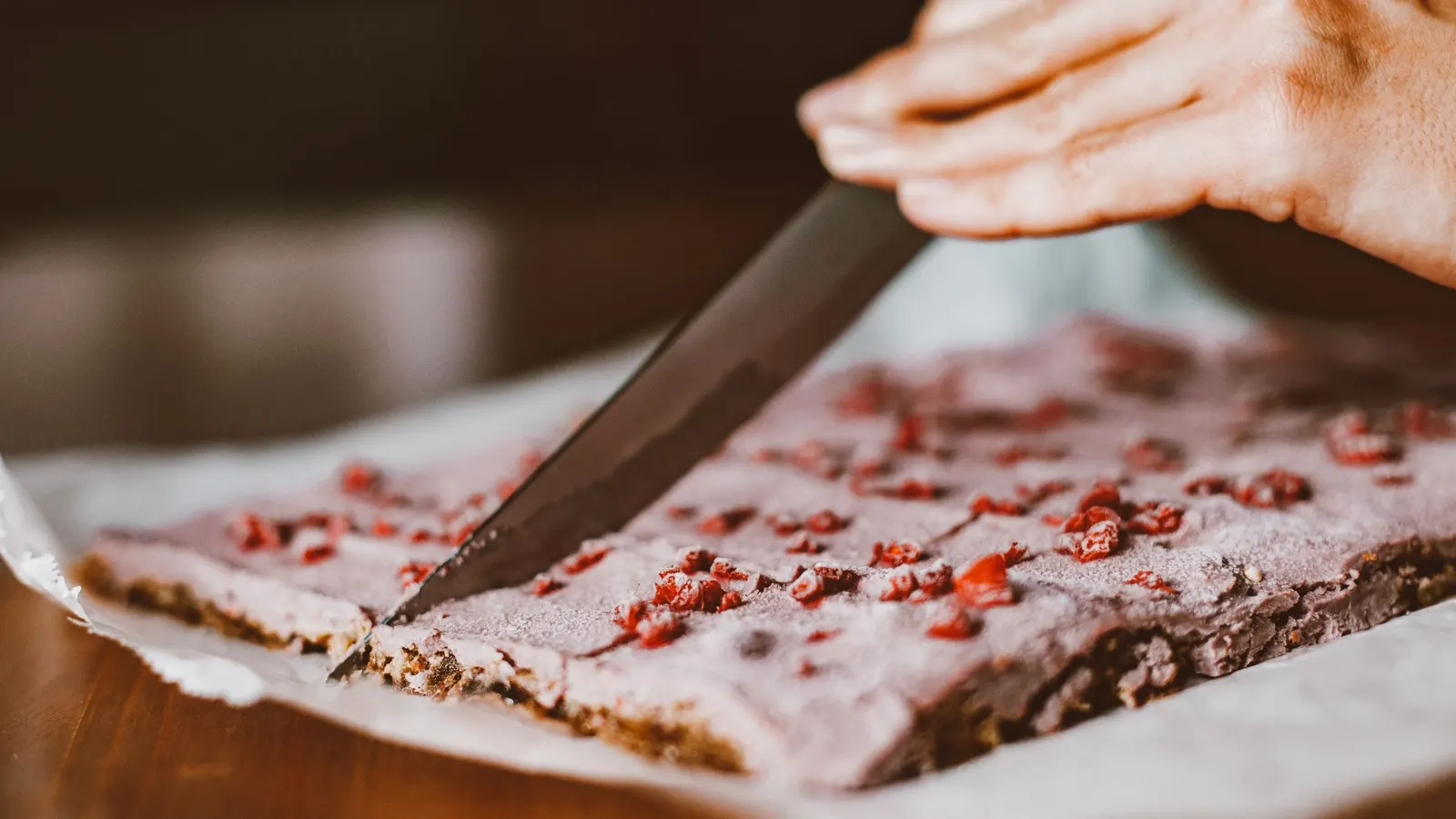Una plancha clara con trozos de fruta roja deshidratada incrustados, cortándose en barras con un cuchillo largo sobre una mesa de madera